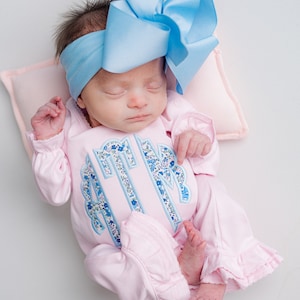 May include: A newborn baby wearing a light pink footed sleeper with a floral monogram and ruffled trim. The baby is also wearing a light blue headband with a large bow. The baby is lying on a pink pillow.