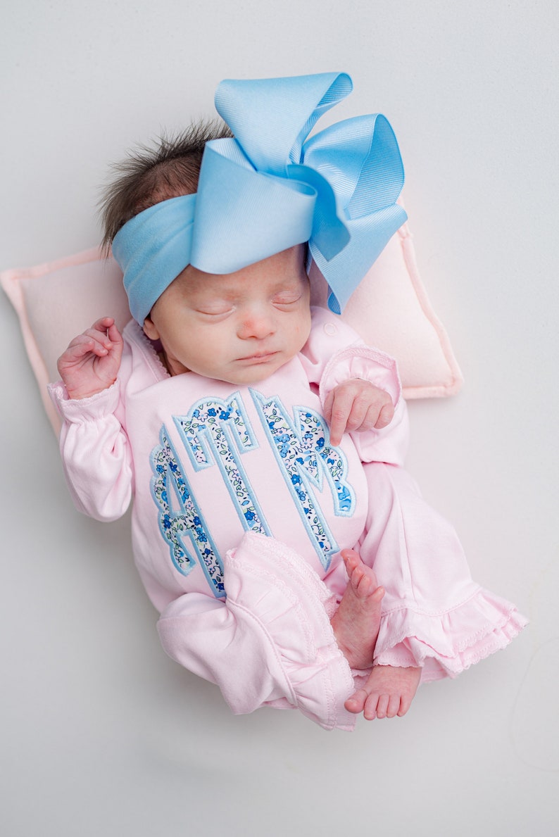 May include: A newborn baby wearing a light pink footed sleeper with a floral monogram and ruffled trim. The baby is also wearing a light blue headband with a large bow. The baby is lying on a pink pillow.