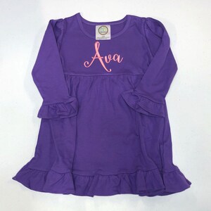 May include: Purple long-sleeved dress with a ruffled hem and sleeves. The dress has a pink embroidered name "Ava" on the front.