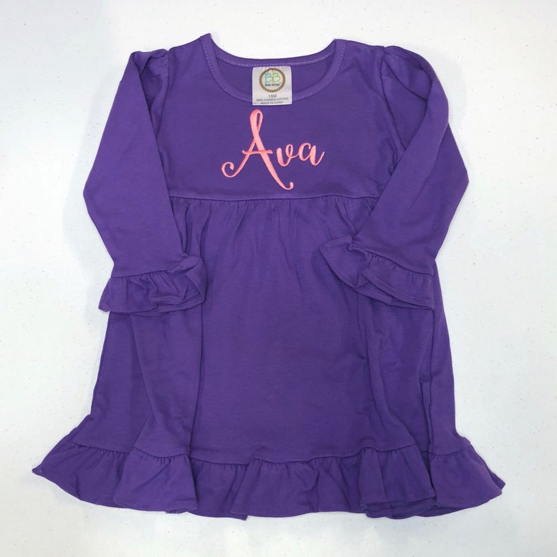 May include: Purple long-sleeved dress with a ruffled hem and sleeves. The dress has a pink embroidered name "Ava" on the front.