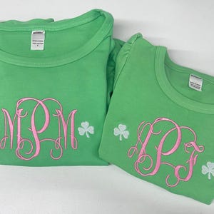 Peut inclure: Deux t-shirts verts avec des monogrammes brodés roses et des accents de trèfles blancs. L'un des t-shirts présente les lettres "MPM", et l'autre affiche "PJ". Les deux t-shirts ont un col rond et sont faits d'une matière douce.