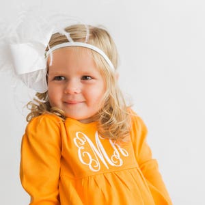 May include: A young girl with blonde hair is wearing a white headband with a large white bow and feathers. She is wearing a bright orange long-sleeved shirt with a white embroidered monogram on the front.