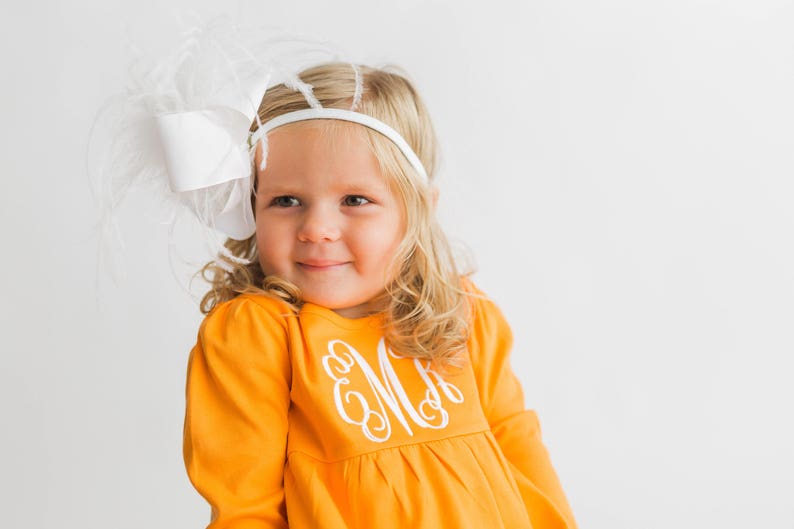 May include: A young girl with blonde hair is wearing a white headband with a large white bow and feathers. She is wearing a bright orange long-sleeved shirt with a white embroidered monogram on the front.