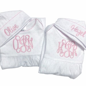 May include: Two white baby gowns with matching hats. The gowns and hats are covered in small pink polka dots. Each gown has a pink monogram and the name "Olive" and "Hazel" embroidered on the hats.
