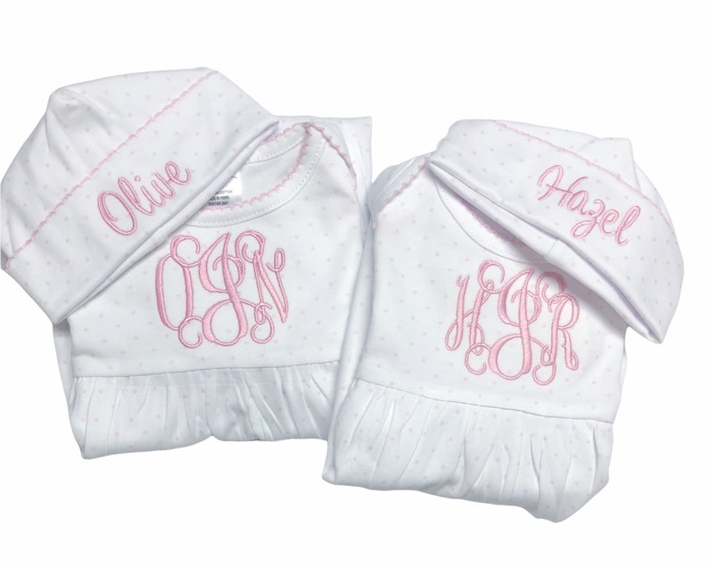 May include: Two white baby gowns with matching hats. The gowns and hats are covered in small pink polka dots. Each gown has a pink monogram and the name "Olive" and "Hazel" embroidered on the hats.