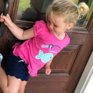 May include: A young girl wearing a pink shirt with a blue and white whale graphic and the word "annie" on it. She is also wearing navy blue shorts and is holding onto a door handle.