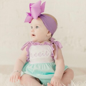 May include: A baby girl wearing a light blue and white ruffle onesie with the name "annie" embroidered on the front. She is wearing a purple headband with a large purple bow on top. The baby is sitting on a white fluffy surface.