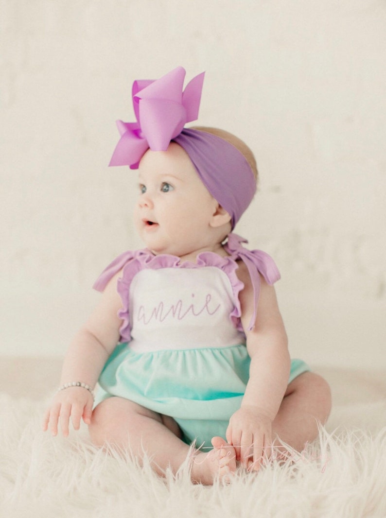 May include: A baby girl wearing a light blue and white ruffle onesie with the name "annie" embroidered on the front. She is wearing a purple headband with a large purple bow on top. The baby is sitting on a white fluffy surface.