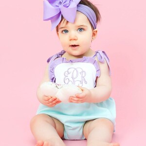May include: A baby girl wearing a light blue and white ruffled romper with a purple bow headband. The romper has a white panel with the monogram "ESR" embroidered in purple.