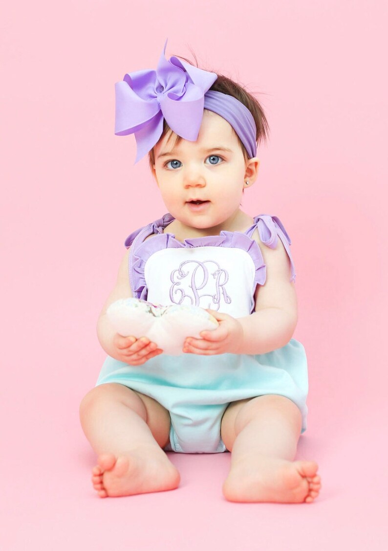May include: A baby girl wearing a light blue and white ruffled romper with a purple bow headband. The romper has a white panel with the monogram "ESR" embroidered in purple.