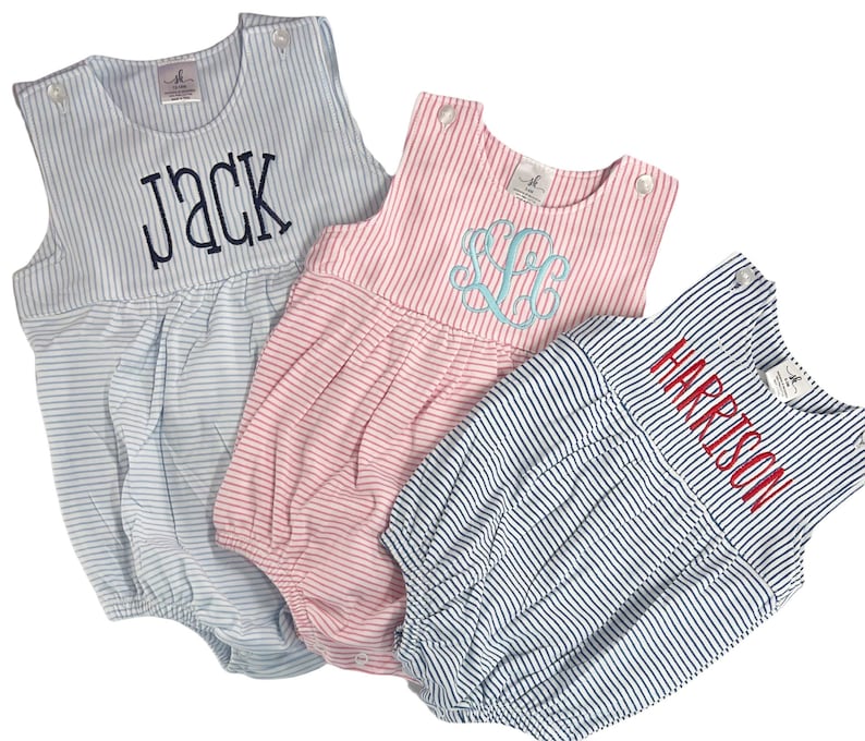May include: Three striped baby rompers in light blue, pink, and navy. Each romper features a different embroidered name or monogram: "JACK", a monogram, and "HARRISON". The rompers have a sleeveless design with button closures at the shoulders.