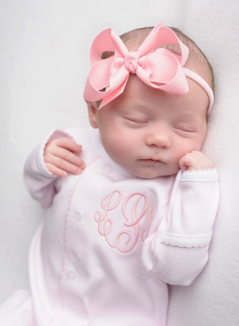 May include: A pink baby onesie with a pink embroidered monogram. The onesie has a white trim around the neckline and sleeves. The baby is wearing a pink bow headband.