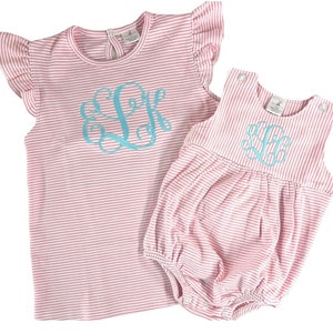 May include: Two-piece set of children's clothing. The set includes a pink and white striped top with ruffled sleeves and a matching romper. Both pieces feature a light blue monogram. The top has a round neckline and the romper has snap closures.