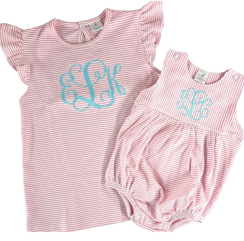 May include: Two-piece set of children's clothing. The set includes a pink and white striped top with ruffled sleeves and a matching romper. Both pieces feature a light blue monogram. The top has a round neckline and the romper has snap closures.