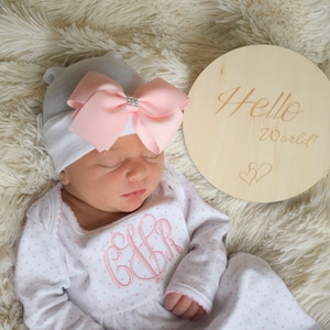 May include: A newborn baby wearing a gray beanie with a pink bow and a white and pink polka dot onesie. A wooden round sign with the words "Hello World!" and two hearts is next to the baby.
