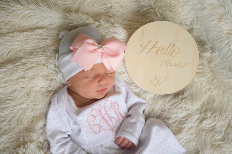 May include: A newborn baby wearing a gray beanie with a pink bow and a white and pink polka dot onesie. A wooden round sign with the words "Hello World!" and two hearts is next to the baby.