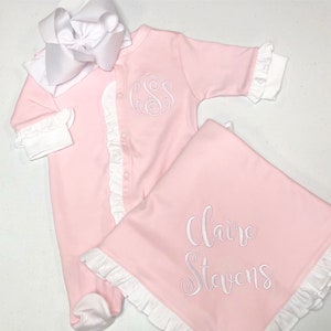 May include: Pink baby outfit with white ruffles and a monogrammed 'SS' on the front. A matching pink blanket with white ruffles and the name 'Claire Stevens' embroidered on it.