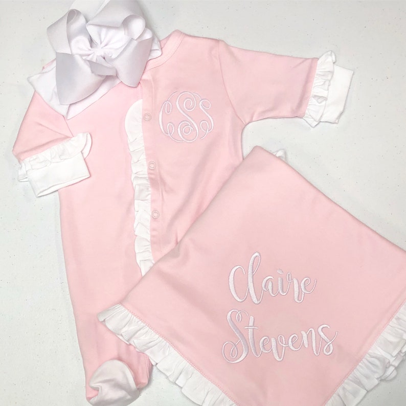 May include: Pink baby outfit with white ruffles and a monogrammed 'SS' on the front. A matching pink blanket with white ruffles and the name 'Claire Stevens' embroidered on it.