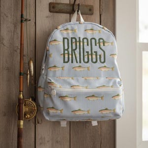 May include: A light blue backpack with a white pinstripe pattern and a repeating fish design. The name "BRIGGS" is embroidered in green on the front. The backpack is hanging on a wooden wall next to a fishing rod.