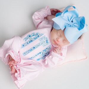 May include: A sleeping infant wearing a pink onesie with a floral monogram and ruffled sleeves. A large light blue bow adorns the baby's head. The baby rests on a small pink pillow.