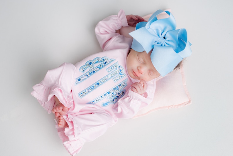 May include: A sleeping infant wearing a pink onesie with a floral monogram and ruffled sleeves. A large light blue bow adorns the baby's head. The baby rests on a small pink pillow.