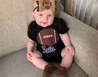 May include: A baby wearing a black bodysuit with a brown football design and the name "Bella" embroidered on it. The baby is also wearing a gold glitter headband with a brown football bow.