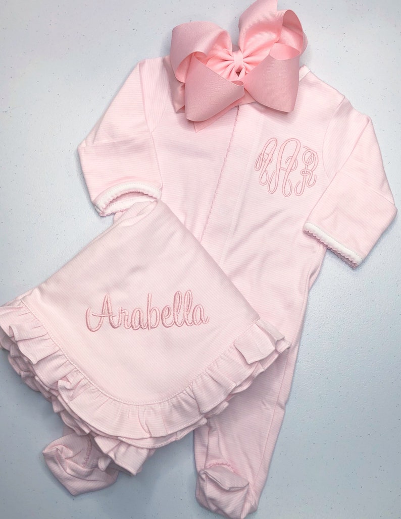 May include: Pink baby outfit with a matching blanket. The outfit has a white trim and is embroidered with the letters "A.A.R." The blanket has a ruffled edge and is embroidered with the name "Arabella".