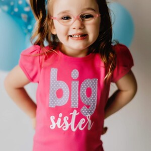K&ouml;nnte beinhalten: Ein junges M&auml;dchen tr&auml;gt eine rosa Brille und ein rosa Shirt mit den Worten "gro&szlig;e Schwester" in grauen und wei&szlig;en Buchstaben. Sie l&auml;chelt und hat ihre Haare zu einem Pferdeschwanz gebunden.