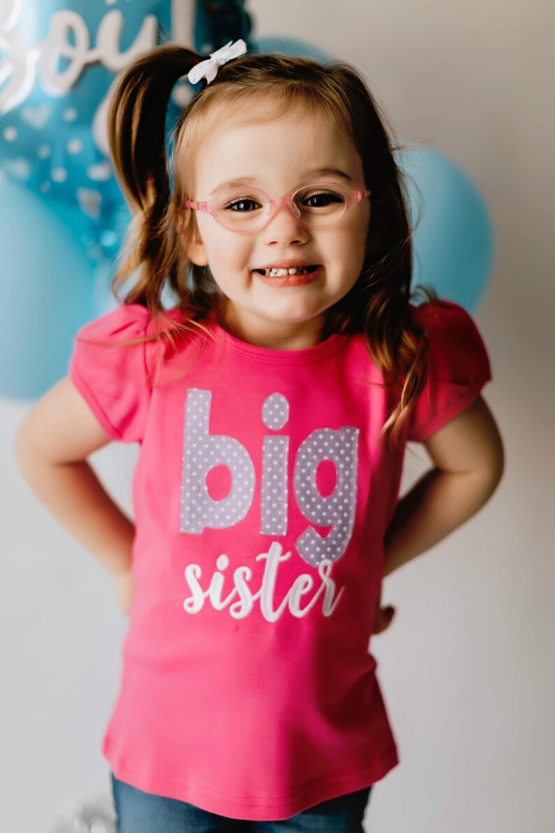K&ouml;nnte beinhalten: Ein junges M&auml;dchen tr&auml;gt eine rosa Brille und ein rosa Shirt mit den Worten "gro&szlig;e Schwester" in grauen und wei&szlig;en Buchstaben. Sie l&auml;chelt und hat ihre Haare zu einem Pferdeschwanz gebunden.