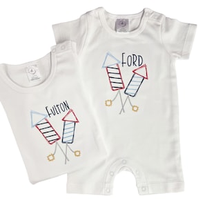 May include: Two white baby outfits, a short-sleeved romper and a vest, each with a colourful embroidered firework design. The romper has the name "FORD" above the fireworks, while the vest says "FULTON". Both outfits have popper fastenings.