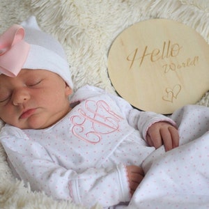 May include: A sleeping infant wearing a white hat with a pink bow and a white onesie with pink polka dots and a pink monogram. A wooden disc with the words "Hello World!" and two hearts is next to the baby.