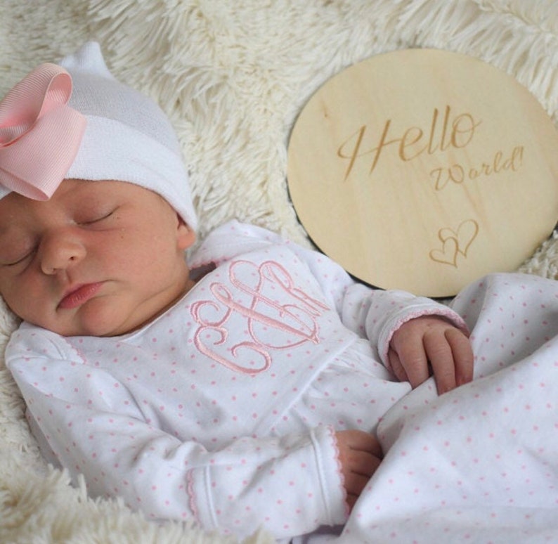 May include: A sleeping infant wearing a white hat with a pink bow and a white onesie with pink polka dots and a pink monogram. A wooden disc with the words "Hello World!" and two hearts is next to the baby.