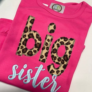 K&ouml;nnte beinhalten: Pinkes T-Shirt mit den Worten "gro&szlig;e Schwester" in hellblauer Schrift. Das Wort "gro&szlig;e" ist in Leopardenmuster.