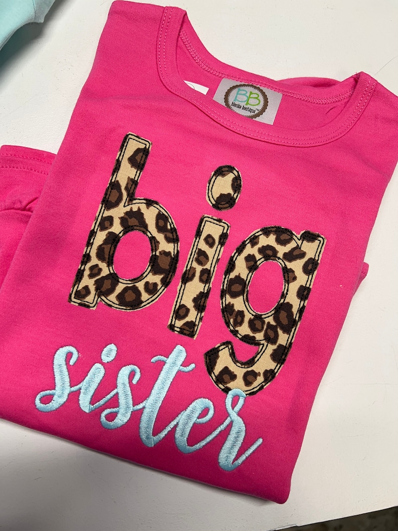 K&ouml;nnte beinhalten: Pinkes T-Shirt mit den Worten "gro&szlig;e Schwester" in hellblauer Schrift. Das Wort "gro&szlig;e" ist in Leopardenmuster.