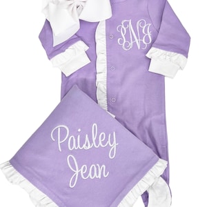 May include: A lavender baby outfit with white ruffles and a white bow. The outfit is monogrammed with the letters "SNB". A matching lavender blanket with white ruffles is embroidered with the name "Paisley Jean".