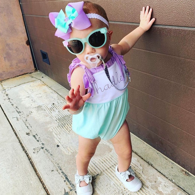 May include: A baby girl wearing a light blue and purple romper with the word "hattie" embroidered on the front. She is wearing a purple and blue bow in her hair, sunglasses, and a pacifier.
