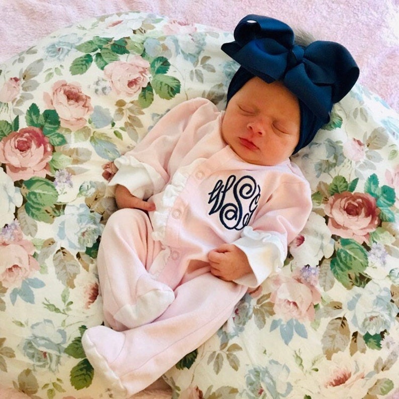 May include: A pink baby onesie with white ruffles and a navy blue monogram on the front. The baby is wearing a navy blue bow headband and is sleeping on a floral patterned pillow.