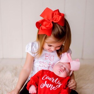 May include: A little girl wearing a white shirt with the text "Big Sister" in pink embroidery is holding a sleeping baby girl. The baby is wearing a red onesie with the text "Courty June" in white embroidery. The older girl is wearing a red bow in her hair and the baby is wearing a pink bow.