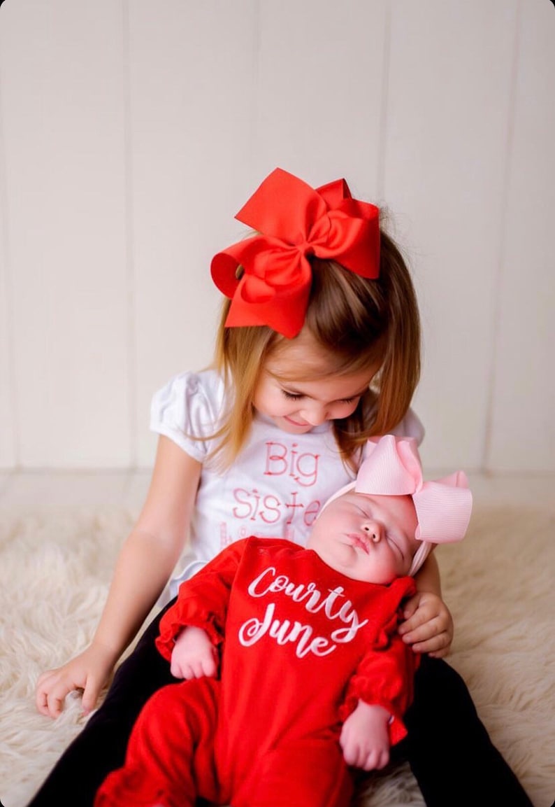 May include: A little girl wearing a white shirt with the text "Big Sister" in pink embroidery is holding a sleeping baby girl. The baby is wearing a red onesie with the text "Courty June" in white embroidery. The older girl is wearing a red bow in her hair and the baby is wearing a pink bow.