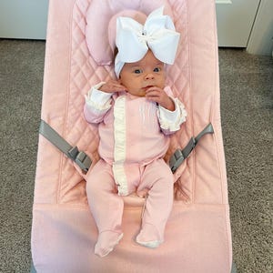May include: A pink baby bouncer with a baby wearing a pink and white outfit and a white bow headband.