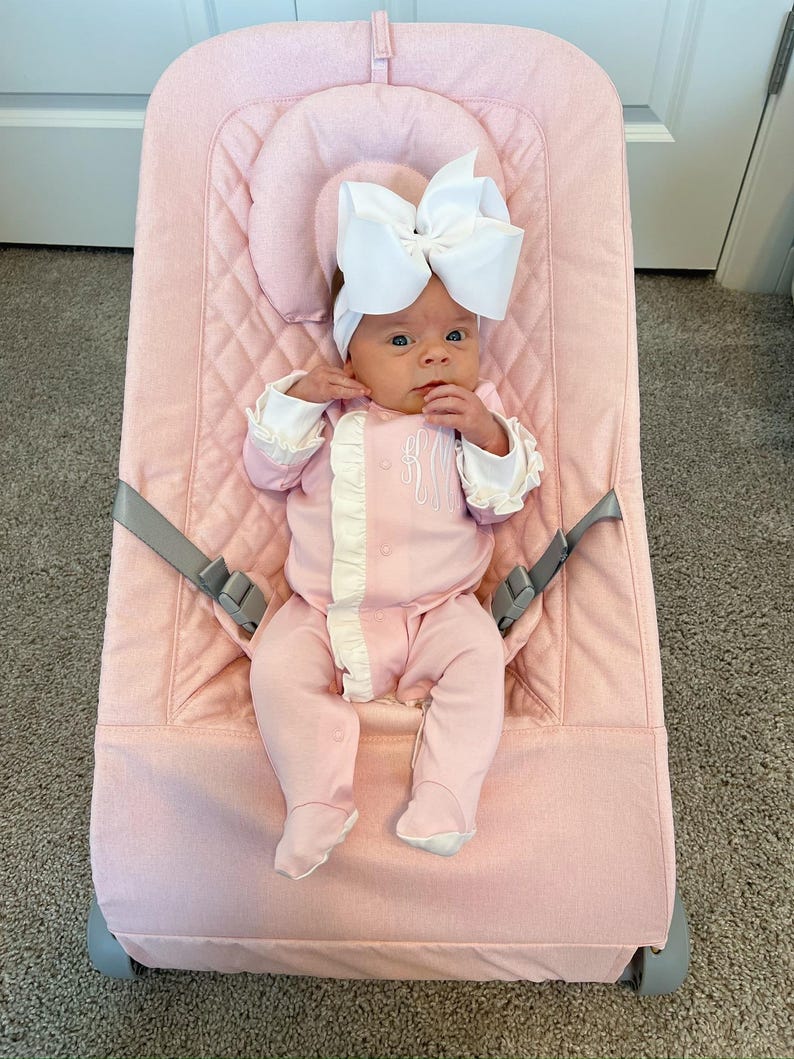 May include: A pink baby bouncer with a baby wearing a pink and white outfit and a white bow headband.