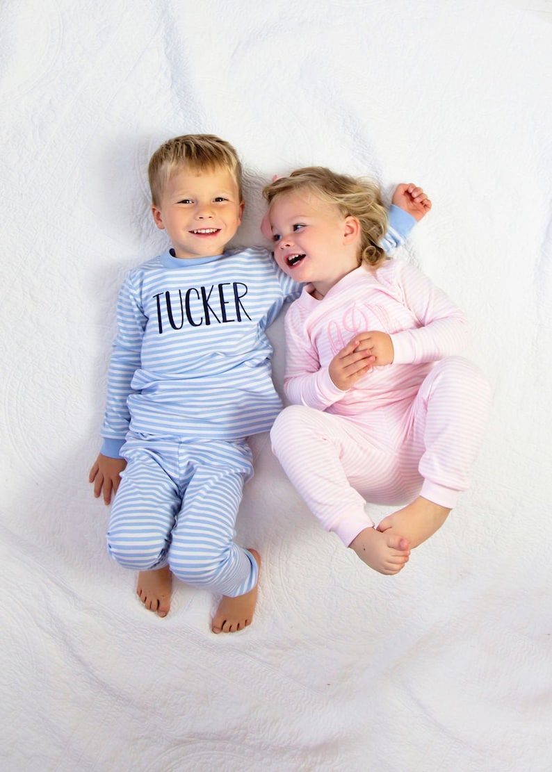Puede incluir: Dos ni&ntilde;os con pijamas a rayas. El ni&ntilde;o lleva un pijama a rayas azul y blanco con el nombre "TUCKER" en la parte delantera. La ni&ntilde;a lleva un pijama a rayas rosa y blanco.