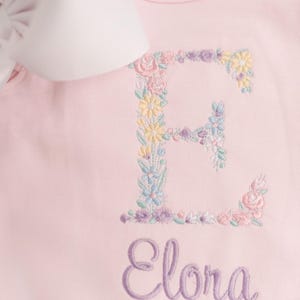 May include: A light pink baby onesie with a floral embroidered letter "E" and the name "Elora" in a cursive font. The floral embroidery includes pastel colors. A white bow is visible in the upper left corner.