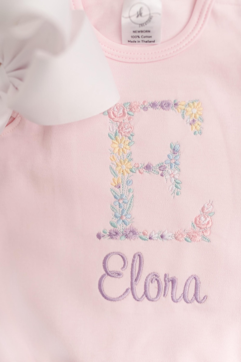 May include: A light pink baby onesie with a floral embroidered letter "E" and the name "Elora" in a cursive font. The floral embroidery includes pastel colors. A white bow is visible in the upper left corner.
