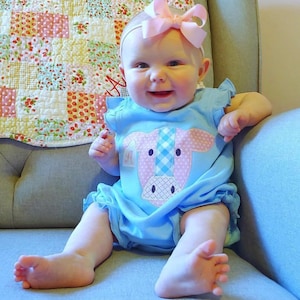 May include: A baby girl wearing a light blue romper with a pink and white cow applique. The romper has a ruffled hem and a pink bow headband.