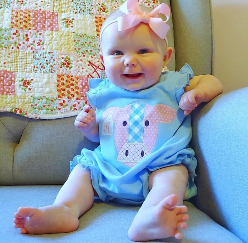 May include: A baby girl wearing a light blue romper with a pink and white cow applique. The romper has a ruffled hem and a pink bow headband.