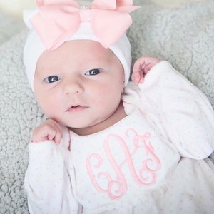 May include: A newborn baby wearing a white hat with a large pink bow and a matching pink monogrammed onesie. The onesie has a bib-like design with the embroidered initials "AJ" in pink. The baby is lying on a soft, gray blanket.