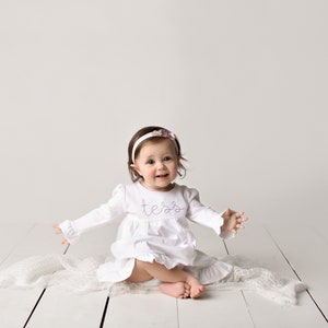 May include: A baby girl wearing a white dress with the name "Tessa" embroidered on the front, sitting on a white blanket with a white headband with a purple flower on it.