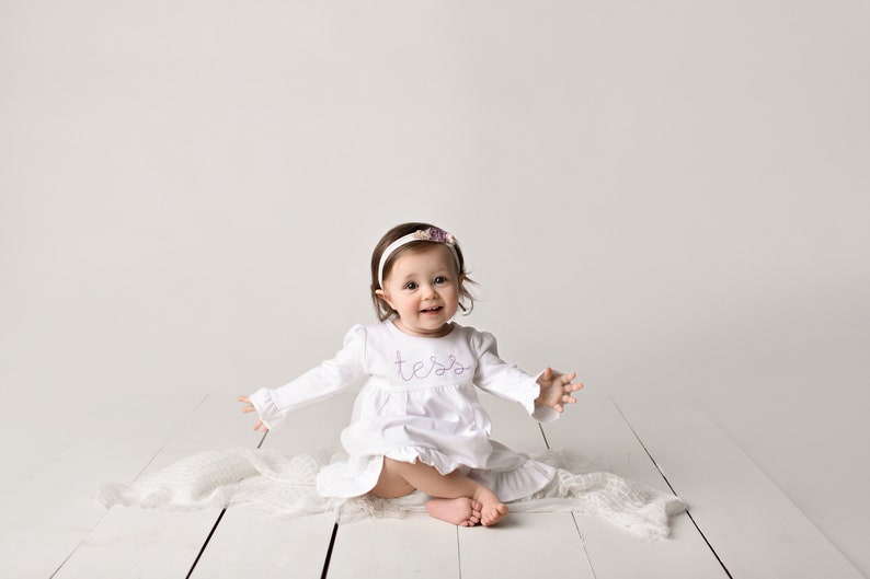 May include: A baby girl wearing a white dress with the name "Tessa" embroidered on the front, sitting on a white blanket with a white headband with a purple flower on it.