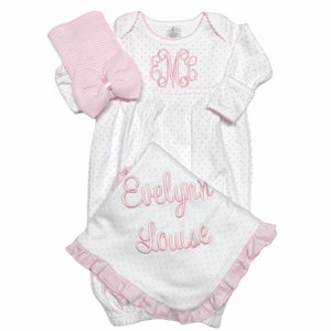 May include: A baby gift set featuring a white and pink polka dot gown, a matching hat with a bow, and a blanket. The gown has a pink embroidered monogram and trim. The blanket is embroidered with the name "Evelynn Louise".